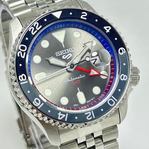Può includere: Un orologio automatico Seiko 5 GMT in acciaio inossidabile. L'orologio presenta un quadrante grigio con indici luminescenti, una lunetta girevole con dettagli blu e neri e una lancetta GMT rossa. Bracciale in acciaio inossidabile.