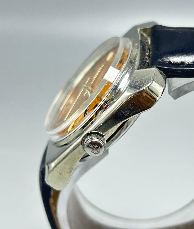May include: A silver-toned wristwatch with a clear, domed crystal and a gold-toned face. The watch has a black leather band and a small, textured crown. The watch is angled, showing the side profile.