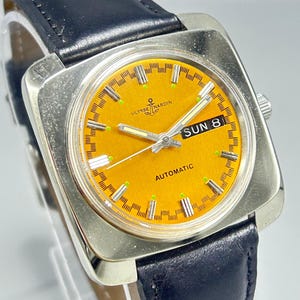 May include: A vintage Ulysse Nardin automatic watch with a square silver case and a black leather band. The watch face is a vibrant orange with silver hands and markers. The day is displayed as "SUN 8".