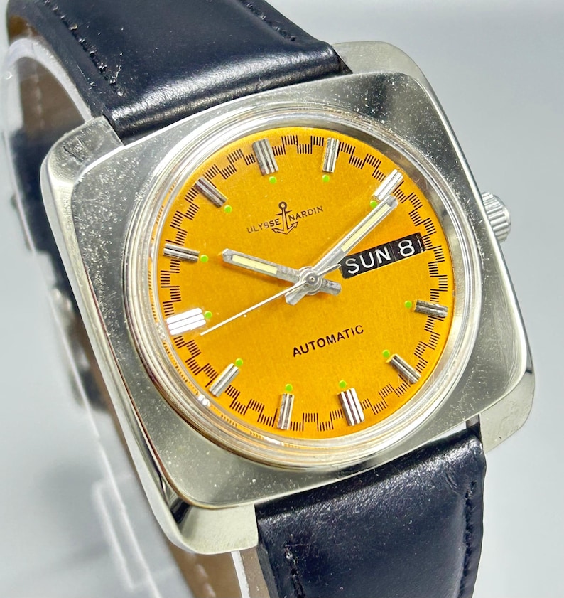 May include: A vintage Ulysse Nardin automatic watch with a square silver case and a black leather band. The watch face is a vibrant orange with silver hands and markers. The day is displayed as "SUN 8".