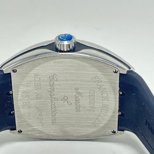May include: A close-up of a luxury watch with a silver case and a dark blue strap. The watch face is rectangular with the words "Master of Complications" and "FRANCK MULLER GENEVE" engraved. The watch has a blue crown.
