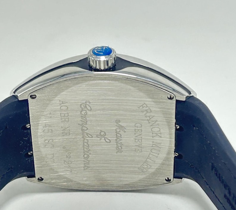 May include: A close-up of a luxury watch with a silver case and a dark blue strap. The watch face is rectangular with the words "Master of Complications" and "FRANCK MULLER GENEVE" engraved. The watch has a blue crown.