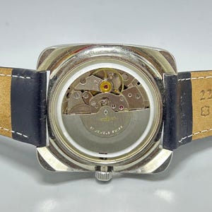 May include: A silver-toned, square-faced wristwatch with a clear back, showcasing the intricate internal mechanisms. The watch has a dark blue leather strap and a silver crown. The back of the watch reads "22 Jewels Swiss".