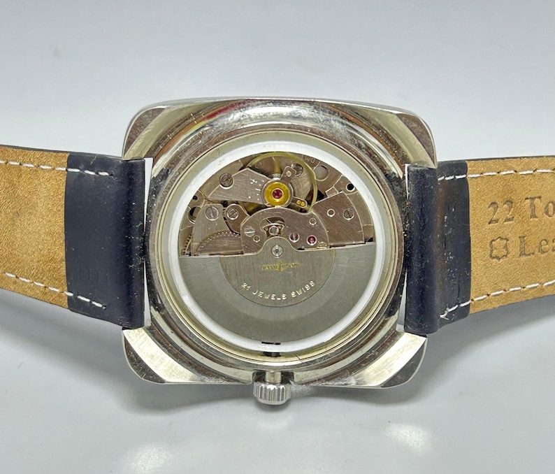 May include: A silver-toned, square-faced wristwatch with a clear back, showcasing the intricate internal mechanisms. The watch has a dark blue leather strap and a silver crown. The back of the watch reads "22 Jewels Swiss".