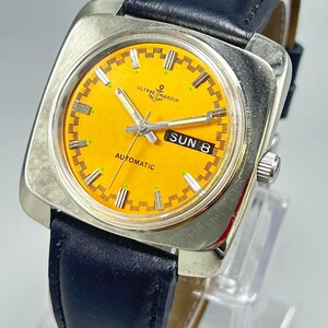 May include: A vintage Ulysse Nardin automatic watch with a square silver case and a bright orange face. The watch features a black leather band and displays the day of the week. The watch face has silver hands and markers.