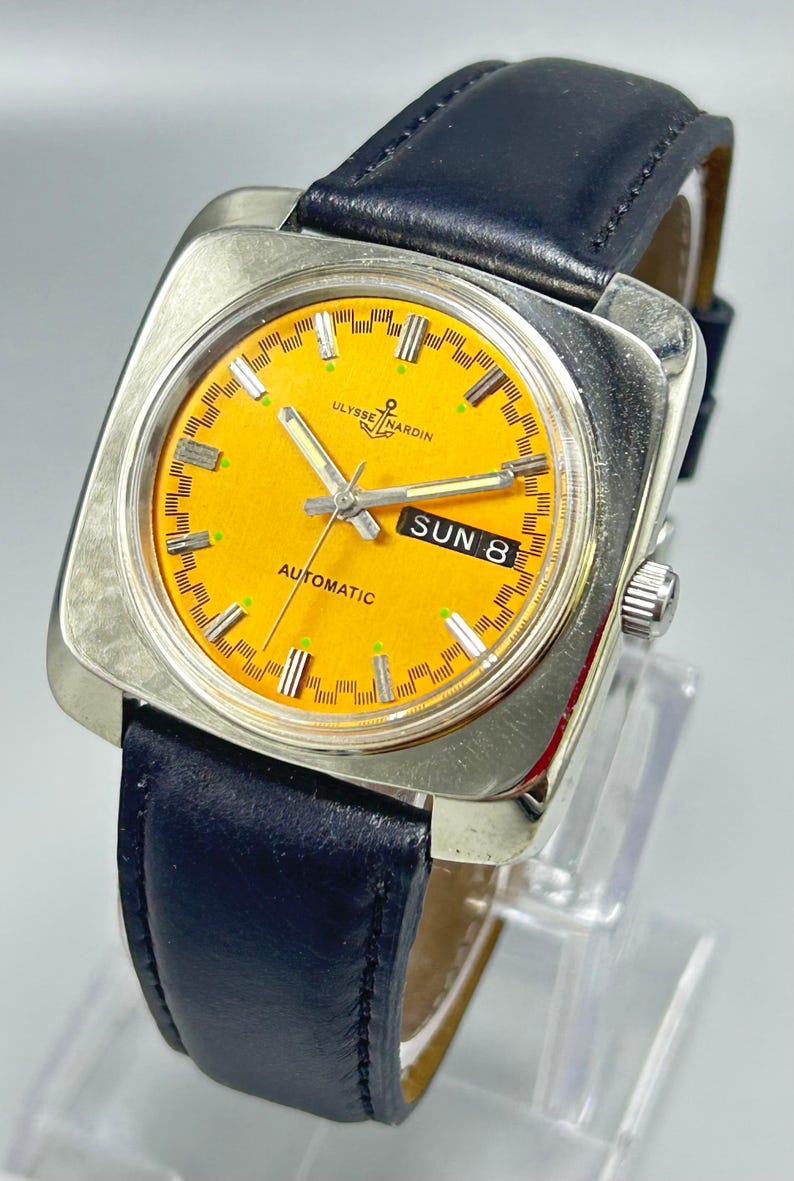 May include: A vintage Ulysse Nardin automatic watch with a square silver case and a bright orange face. The watch features a black leather band and displays the day of the week. The watch face has silver hands and markers.
