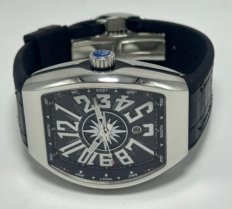 May include: A silver wristwatch with a black face, large white numerals, and a compass design. The watch has a black rubber strap and a silver bezel. The watch face displays the words "EAST", "SOUTH", "WEST", and "NORTH".