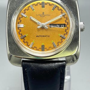 May include: A vintage Ulysse Nardin automatic wristwatch with a square silver case and a black leather band. The watch face is orange with silver hands and markers, and displays "SUN 8". The watch has the brand name "Ulysse Nardin" on the face.