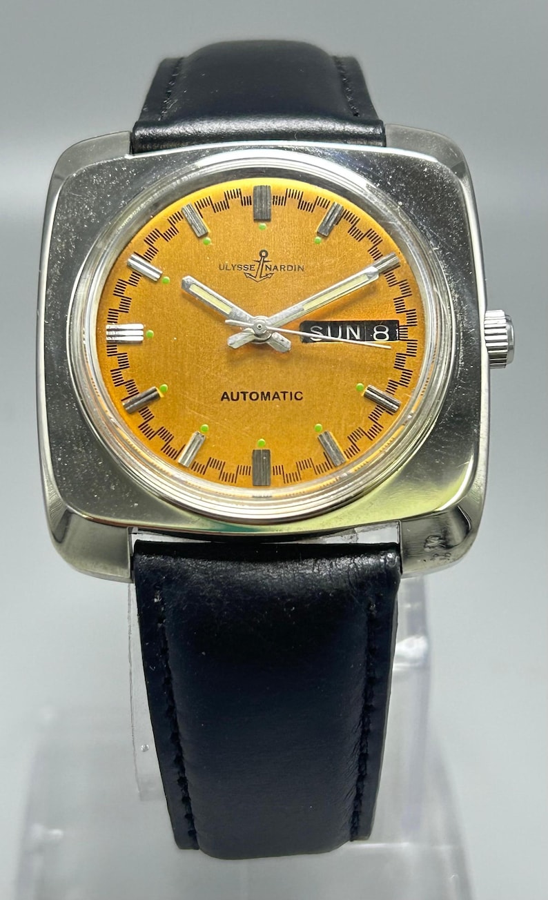 May include: A vintage Ulysse Nardin automatic wristwatch with a square silver case and a black leather band. The watch face is orange with silver hands and markers, and displays "SUN 8". The watch has the brand name "Ulysse Nardin" on the face.