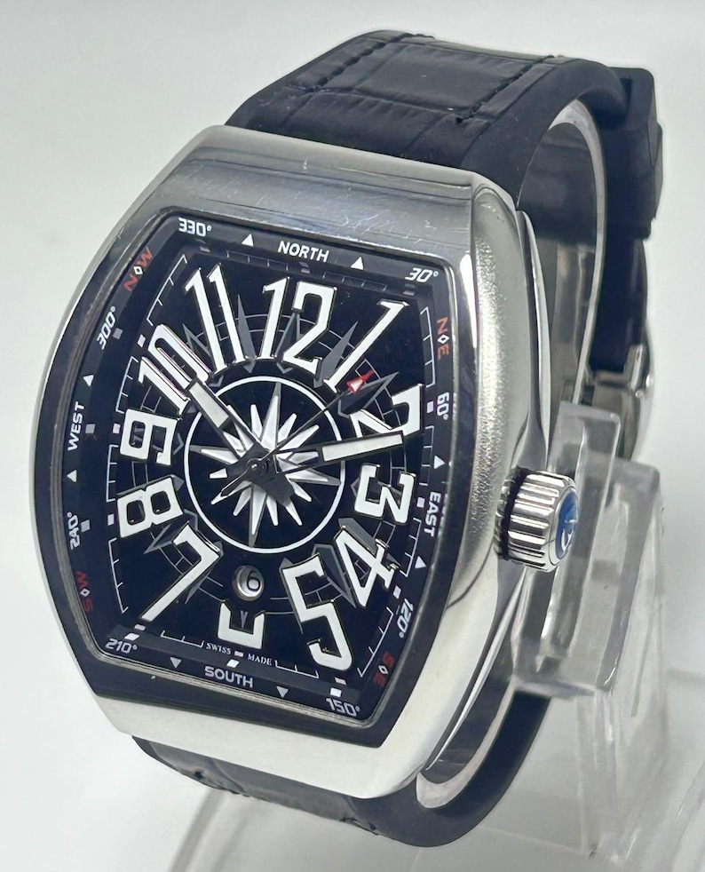 May include: A silver wristwatch with a black face and a black leather strap. The watch face displays large white numerals, a compass design, and the words "SWISS MADE". The watch has a rectangular case and a crown with a blue stone.