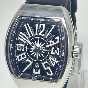 May include: A silver wristwatch with a black face and a black leather strap. The watch face displays large white numerals, a compass rose design, and the words "SWISS MADE". The watch has a rectangular case and a crown with a blue gem.
