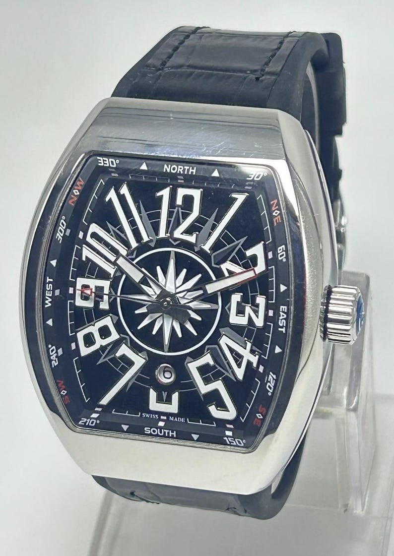 May include: A silver wristwatch with a black face and a black leather strap. The watch face displays large white numerals, a compass rose design, and the words "SWISS MADE". The watch has a rectangular case and a crown with a blue gem.