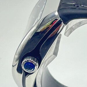 May include: Close-up of a silver wristwatch with a black leather strap. The watch face has a blue accent and a silver bezel. The watch is displayed on a white background.