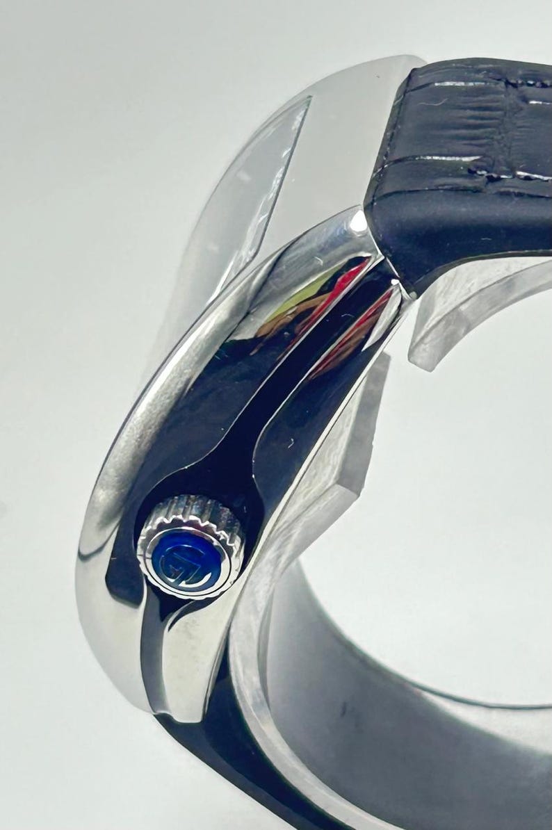 May include: Close-up of a silver wristwatch with a black leather strap. The watch face has a blue accent and a silver bezel. The watch is displayed on a white background.