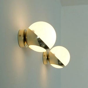 May include: Two modern wall sconces with a gold finish and spherical, frosted glass shades. Each sconce has a diagonal gold accent. The sconces are mounted on a light-colored wall, providing ambient lighting.
