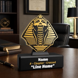 May include: A black and gold desk award featuring a stylized pharaoh mask emblem mounted on a rectangular base. The base displays the text Name, # - Chapter - Crossed, and Line Name in white and gold lettering against a solid black background.