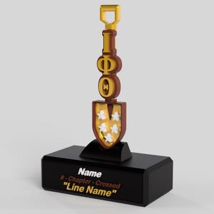 May include: A black trophy with a shovel design. The shovel is brown and gold, with the letters "IΦO" in gold. The shovel blade has gold stars. The base is black with the text "Name", "# - Chapter - Crossed", and "Line Name".