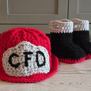 May include: A red crocheted hat with a light gray patch featuring the black embroidered letters CFD, accompanied by a pair of black crocheted booties with light gray cuffs and red soles, all crafted with a thick yarn texture.