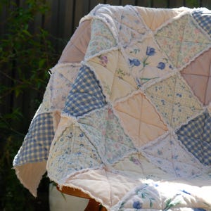 May include: A patchwork quilt in pastel shades, draped over a wooden chair. The quilt is made from various fabrics, including pink, blue gingham, and floral prints. The edges are finished with a ruffled trim, giving it a cosy, vintage appearance.