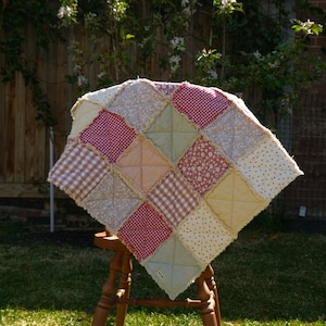 May include: A square rag quilt featuring a patchwork design of various fabric squares in shades of red, white, yellow, and sage green. The quilt displays a mix of gingham, floral, and polka dot patterns with exposed, frayed seams in a cross stitch.