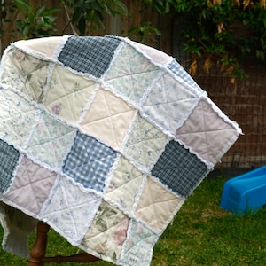 May include: A patchwork quilt featuring a mix of floral, gingham, and solid fabric squares in pastel shades. The quilt is displayed on a wooden chair outdoors, highlighting its soft texture and design. The quilt measures approximately 100 cm by 100 cm.