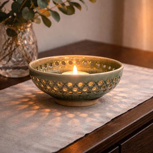 May include: A green ceramic candle holder with a tea light candle. The bowl-shaped holder has a perforated design and sits on a light-colored table runner. A glass vase with greenery is in the background.