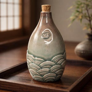 May include: A ceramic bottle with a cork stopper. The bottle has a brown and green gradient with a white swirl design. The bottom of the bottle features a wave pattern. The bottle is sitting on a wooden tray.