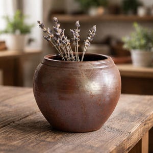 May include: A round, brown ceramic vase with a wide opening, filled with dried lavender stems. The vase has a rustic, textured finish and sits on a wooden table. The background is blurred, suggesting a home decor setting.