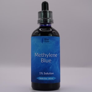 Puede incluir: Una botella de vidrio azul oscuro con un tapón cuentagotas negro. La etiqueta dice "Methylene Blue 1% Solution" y "Bottle Size: 100 ml". La botella está llena de un líquido azul oscuro. La etiqueta también presenta el texto "Blue Gum Solutions".