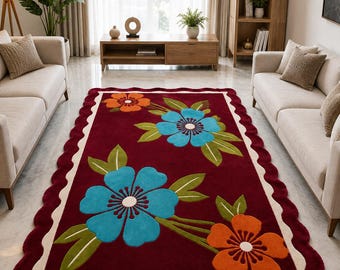 Puede incluir: Una alfombra rectangular con fondo color burdeos intenso que presenta grandes motivos florales en azul y naranja vibrantes con hojas verdes. La alfombra tiene un borde festoneado de color crema y una textura suave y acolchada.