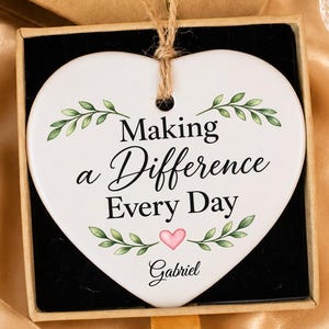 May include: A white heart-shaped ceramic ornament with green leaf illustrations and the black text "Making a Difference Every Day" above a small pink heart and the name "Gabriel," presented in a brown cardboard gift box with a twine hanger.