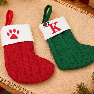 May include: Two cable-knit Christmas stockings with white cuffs. The red stocking features a red paw print on the cuff, while the green stocking displays a red letter K with a small Santa hat icon embroidered on the white cuff, each with a loop.