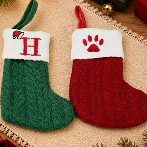 May include: Two knit Christmas stockings with hanging loops. One is green with a white cuff featuring a red letter H and a small Santa hat. The other is red with a white cuff featuring a red paw print. Both have a cable knit texture.