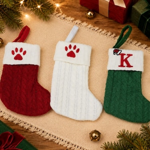 May include: Three cable-knit Christmas stockings with hanging loops. The red stocking features a white cuff with a red paw print. The white stocking has a white cuff with a red paw print. The green stocking has a white cuff with a red letter K.