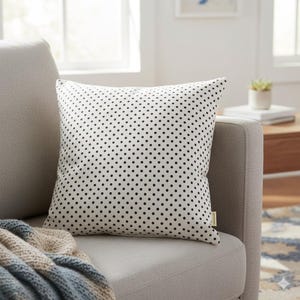 May include: Square throw pillow with a white background and a pattern of evenly spaced black polka dots. The pillow is on a gray sofa, with a blue and tan blanket visible. A small tag is sewn on the pillow's corner.