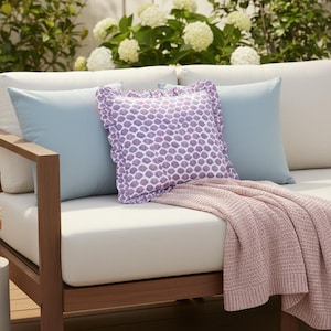 以下が含まれることがあります： ライトブルーと紫の模様のクッション、ピンクのニットスローが付いた屋外用ソファ。ソファは木製のフレームとクリーム色のクッションです。小さな鉢植えの植物がサイドテーブルに置かれています。