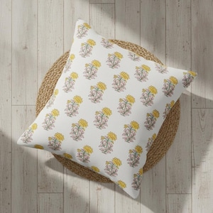 May include: Square white cushion featuring a repeating floral design. The flowers are yellow with pink and green details. The cushion is placed on a round, woven, tan mat, set against a light wood floor.
