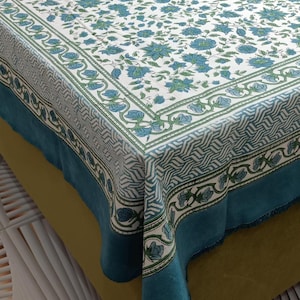 May include: A white tablecloth with a blue floral pattern and a teal border. The floral design features blue flowers and green leaves. The border has a geometric pattern and a fringe edge. The tablecloth is draped over a bed.