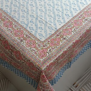 May include: A white tablecloth with a repeating blue pattern and a pink floral border. The border features intricate floral designs in pink, yellow, and green. The tablecloth's edge has a blue and white checkered pattern.