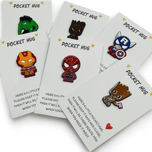 May include: A collection of six enamel pins featuring various comic book characters, including Hulk, Groot, Captain America, Iron Man, and Spider-Man. Each pin is attached to a white card labelled POCKET HUG with printed text about keeping it close.