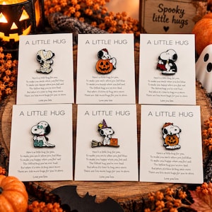 May include: Six enamel pins featuring Snoopy in various Halloween costumes, including a mummy, a pumpkin, a vampire, a ghost, a witch, and a pumpkin carrier. Each pin is mounted on a white card titled A LITTLE HUG with printed poem text.