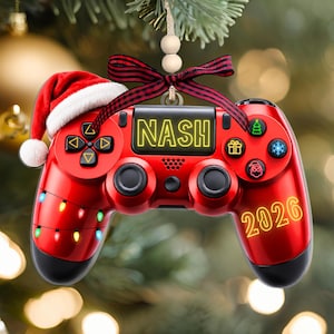 May include: A red video game controller ornament featuring a Santa hat, a black and red plaid ribbon, and two wooden beads. The controller displays the name NASH, the year 2026, and icons of a Christmas tree, gift, and Santa Claus with string lights.