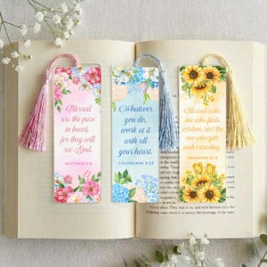 Könnte beinhalten: Drei rechteckige Lesezeichen aus Papier mit Quasten auf einem aufgeschlagenen Buch. Das linke Lesezeichen ist rosa mit Blumen und dem Text Blessed are the pure in heart, for they will see God. Das mittlere ist blau mit Blumen und dem Text Whatever you do, work at it with all your heart. Das rechte ist gelb mit Sonnenblumen und dem Text Blessed is the one who finds wisdom, and the one who gets understanding.