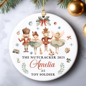 Könnte beinhalten: Weißer Keramik-Weihnachtsschmuck mit einer Aquarell-Illustration von Figuren aus dem Nussknacker-Ballett. Der Schmuck enthält den Text "THE NUTCRACKER 2025", "Amelia" und "AS TOY SOLDIER". Der Schmuck ist mit festlichen Elementen verziert.