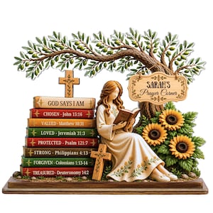 May include: A decorative figurine featuring a person reading a book under an olive tree. A stack of books with religious text sits beside them, topped with a wooden cross. A hanging sign reads Sarah's Prayer Corner, with sunflowers nearby.