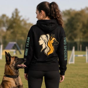 Puede incluir: Una sudadera negra con capucha que presenta un gráfico en la espalda con el perfil de una mujer y el perfil de un perro mirando en direcciones opuestas. El texto ONE TEAM está impreso en la manga izquierda y TWO SPECIES en la derecha en verde claro.