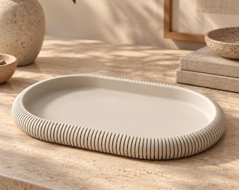 Puede incluir: Una bandeja de forma ovalada con un acabado mate en color blanco roto. La bandeja presenta una base interior lisa y plana rodeada por un borde grueso y elevado con una textura vertical acanalada que recorre todo el borde exterior.