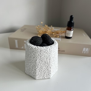 May include: A white, textured, hexagonal ceramic pot filled with black, porous stones. A small brown bottle with a dropper is to the right. A book and dried flowers are in the background. The pot is on a white surface.