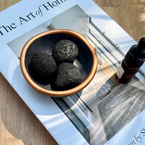 May include: A small, dark blue bowl with a copper-coloured rim holds three black, porous lava rocks. A small amber glass bottle with a dropper is next to the bowl. The items rest on a magazine titled "The Art of Home."
