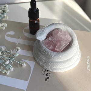 May include: A white ceramic snake-shaped bowl filled with pink rose quartz crystals. A small brown glass bottle with a black dropper sits beside the bowl. The items are on a light-coloured surface with text.
